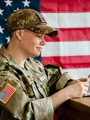 A soldier in camouflage uniform writes on a notebook at a wooden desk, with a large American flag in the background, symbolizing duty and patriotism.