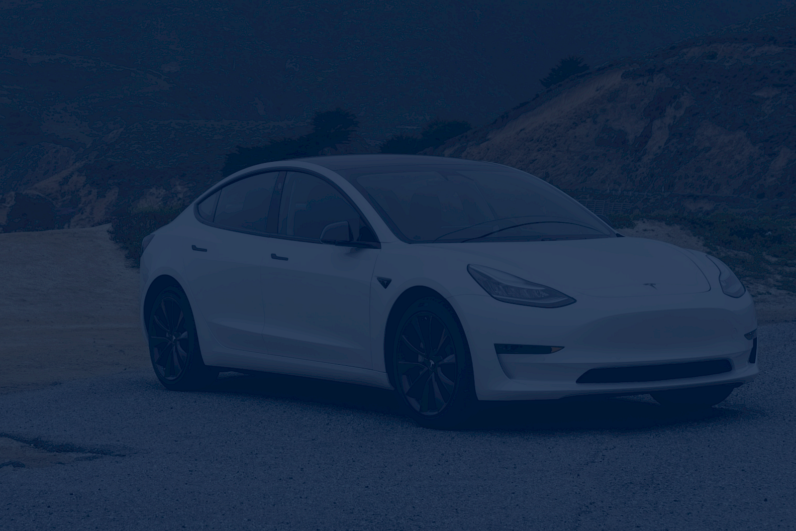 A white Tesla Model Y parked on a coastal road, with hills in the background, under a blue-tinted overlay.