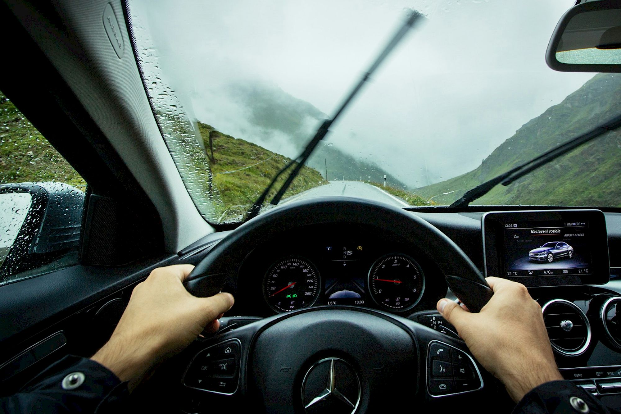 A driver&rsquo;s view from inside a car on a rainy mountain road, steering wheel centered, windshield wipers in motion, dashboard and infotainment screen ahead.