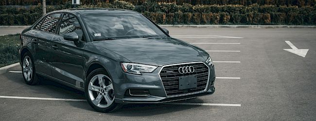A sleek dark gray Audi sedan parked diagonally in a nearly empty parking lot, front end facing the camera, with white-lined spaces and trees in the background.
